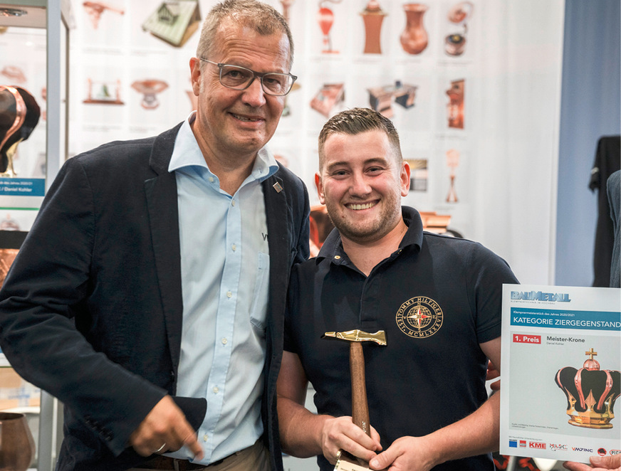 Daniel Kohler setzt der Veranstaltung die Krone auf. Seine Meisterkrone zierte während der Dach + Holz International 2022 den BAUMETALL-Messestand. Zum ersten Preis in der Kategorie „Bester Ziergegenstand“ gratuliert ihm Uwe Nagel von VMZinc mit einer Spezialschulung