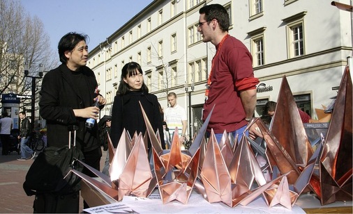 Japanische Touristen erfahren bei der Kranichaktion der Robert-Mayer-Schule in der Stuttgarter Fußgängerzone, dass Origamikraniche aus Metall gefaltet werden können, und sind begeistert