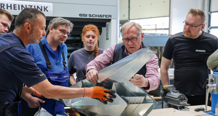 Gerne! Hier ist ein präziser, themenrelevanter und zielgruppengerechter Alternativtext für das beschriebene Bild: Alt-Text: "Erfahrener Metallbaumeister demonstriert einer Gruppe von Fachhandwerkern die Verarbeitungstechnik an einer komplexen Fassadenkonstruktion aus Aluminium in einer Werkstattumgebung. - © Bild: BAUMETALL