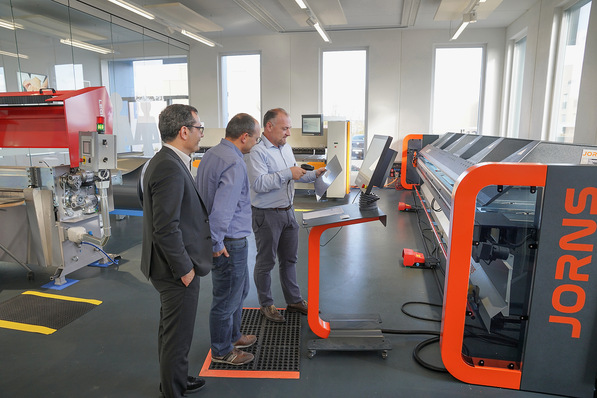  Andrea Gervasio (Spiegel) scannt den QR-Code zur Maschineneinstellung auf einer JORNS-Langabkantmaschine im Beisein von Luigi Greco (Gesacon) und Roger Lehner (Alurex)   ﻿