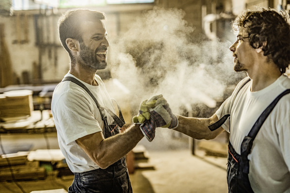 Happy male carpenters greeting each other while holding hands in a workshop.