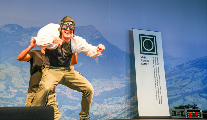 Zwei Personen treten auf der Bühne auf, eine davon trägt eine Pilotenbrille und einen Helm und ahmt das Fliegen mit Bergen im Hintergrund nach. - © Bild: BAUMETALL